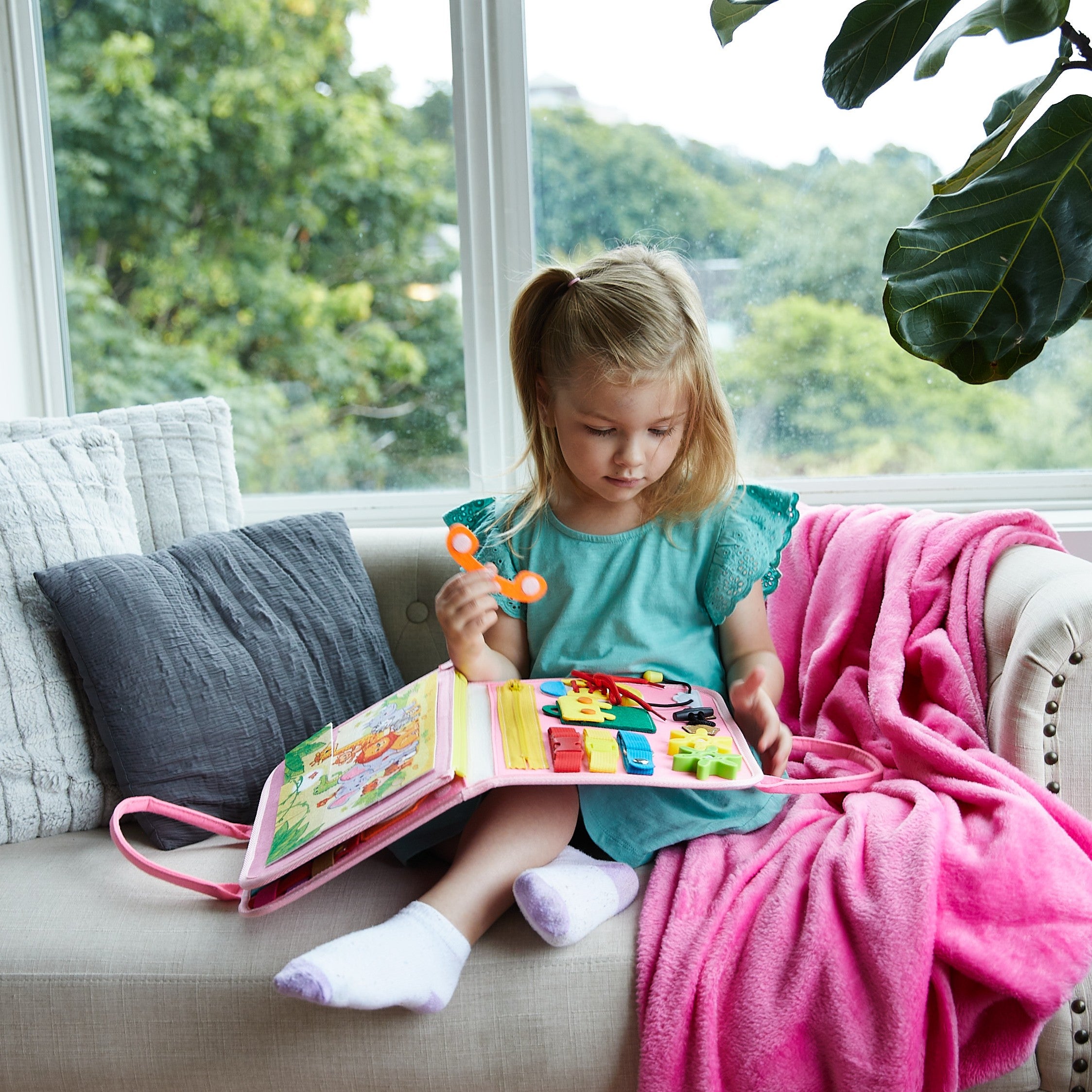 Child playing with a colorful toy book on a couch with a window and greenery in the background