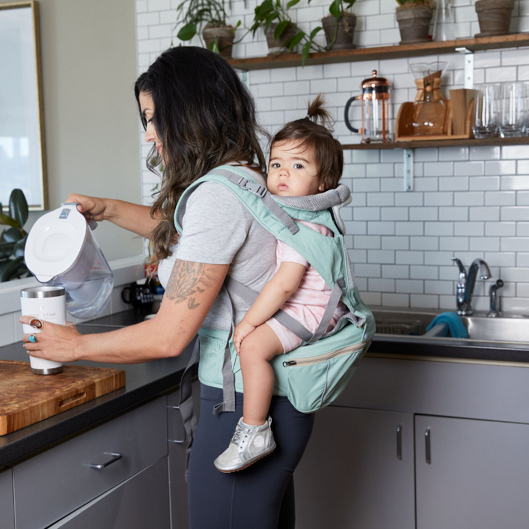 Woman in a kitchen using a water filter while holding a baby in a carrier.